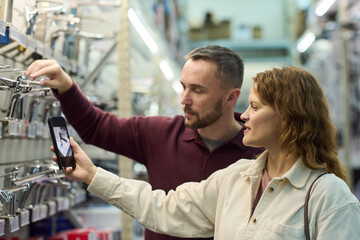 Caucasian man and Caucasian woman examining faucets in hardware store, woman holding smartphone and taking photo while man reaching for display, both focused on selection