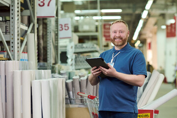 Portrait of man standing in hardware store holding digital tablet and smiling, surrounded by rolls of material and industrial shelving, wearing employee badge