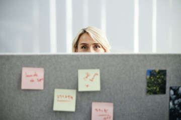 Caucasian young adult woman peeking over office partition, only eyes and forehead visible, standing behind gray divider with colorful sticky notes attached, modern workspace setting