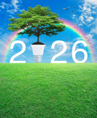 Big green tree on soil with light bulb and 2026 letter on green grass field over rainbow, birds and blue sky with white clouds, Happy new year 2026 green ecology and saving energy concept