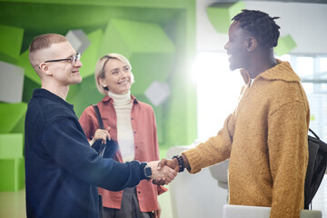 Caucasian young adult man shaking hands with Black young adult man while smiling, standing in...