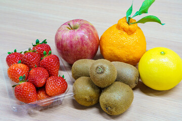 Assortment of fresh fruits on a wooden table. Strawberries in a plastic pack, red apple, kiwi fruits, Shiranui citrus and yellow orange. Healthy dessert ingredients.