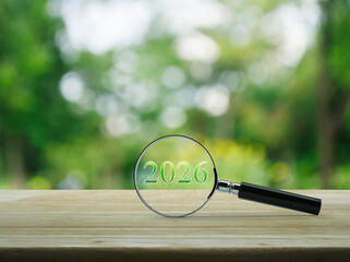 Magnifying glass with 2026 text and financial graph on wooden table over blur green tree in garden, Business happy new year 2026 research concept