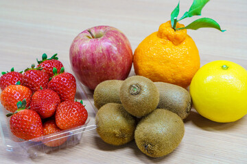 Assortment of fresh fruits on a wooden table. Strawberries in a plastic pack, red apple, kiwi fruits, Shiranui citrus and yellow orange. Healthy dessert ingredients.