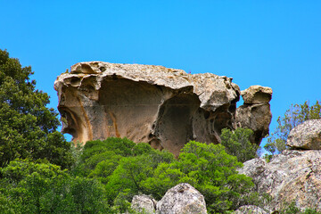Dramatic geological scenery showing weathered granite stones with deep cavities surrounded by green mediterranean vegetation under a bright blue sky