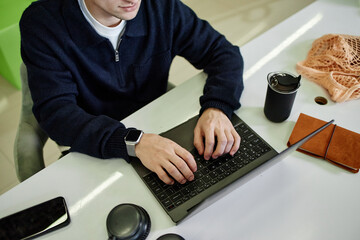 Young adult Caucasian man working on laptop at desk, hands typing on keyboard, wearing smartwatch, reusable cup and notebook nearby, partial face visible, casual workspace setting