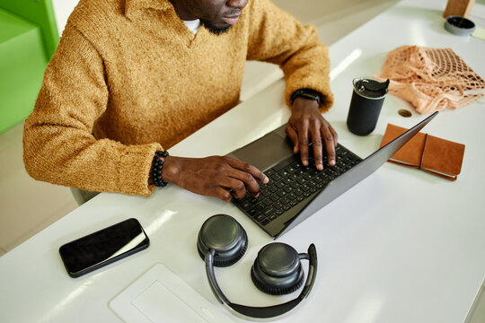 Young adult Black man working on laptop at desk, hands typing on keyboard, smartphone and wireless headphones nearby, reusable cup and notebook on workspace, partial face visible