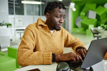 Young adult Black man working on laptop in modern office environment, focusing on screen while typing, sitting at desk with blurred green geometric background visible behind