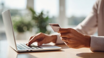 Hands are holding a credit card while typing on a laptop keyboard. The setting features natural daylight in a clean workspace with no distractions
