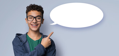 Happy smiling black curly hair man wear braces brackets, glasses, hoodie, advertise pointing at white empty mockup speech bubble, isolated gray grey background. Dental care, ophthalmology ad