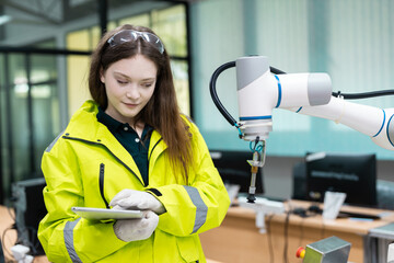 Female engineer using digital tablet to control or monitor robotic arm in modern industrial facility, representing smart manufacturing and automation technology.