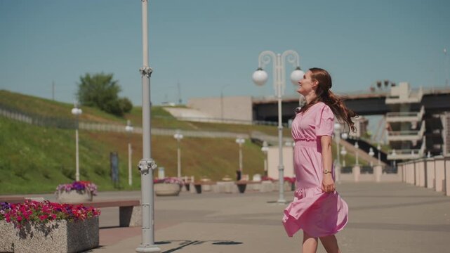 young woman in pink dress strolling along sunlit promenade, braided hair catching breeze, smiling gaze upward, light footsteps past lamp posts and flower planters, green embankment and modern urban