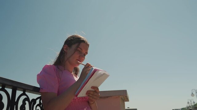 young white woman writing in notebook on rooftop under clear sky focused expression, pen in hand, checking notes, drafting script and planner entries, journalist and editor roles suggested