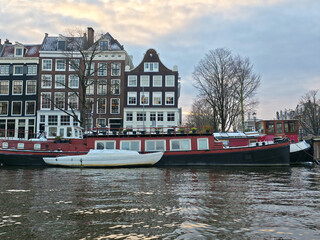 Two houseboats moored on a tranquil Amsterdam canal, featuring the city's signature narrow, gabled buildings lining the snowy bank under an overcast sky