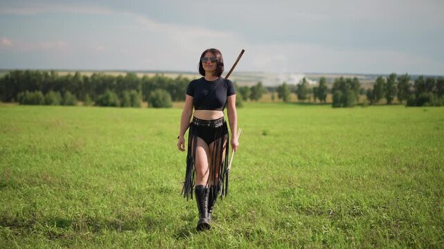 caucasian female warrior walking toward camera confident frontline ranger advancing through open meadow spear over shoulder leather fringe outfit steady gaze into lens purposeful stride bright