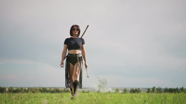caucasian female warrior walking across grass, leather outfit and boots, staff slung over shoulder, solitary march toward distant horizon under moody sky, faint dust plume in background, cinematic