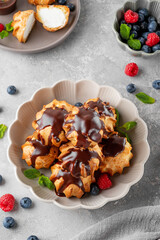 Profiteroles with vanilla cream, fresh berries and chocolate glazed in a bowl on a gray concrete background. A delicious French dessert.