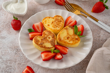 Heart-shaped cottage cheese pancakes or syrnyky with sour cream and fresh strawberries on a plate. A delicious breakfast for Valentine's Day or Mother's Day. Top view, copy space.
