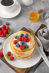 A stack of pancakes with whipped cream, fresh berries and honey on a white plate on a gray concrete background.