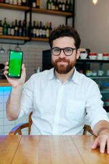 Portrait of Caucasian young adult man with beard and glasses sitting at table holding smartphone with green screen in hand looking into camera in modern cafe setting