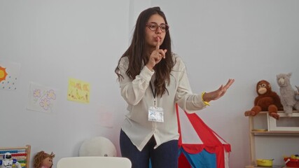 Woman teaching in a lively kindergarten setting with colorful toys and drawings, wearing glasses and a badge, emphasizing a nurturing educational environment.