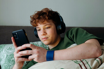 Teenage Caucasian boy lying on bed wearing headphones using smartphone holding device with both hands focused on screen relaxing in bedroom technology leisure concept