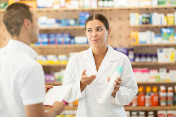 Female consultant pharmacist holds bottle of shampoo and offers to man client to purchase remedy to...