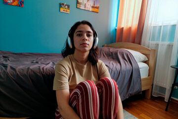 Portrait of young adult Hispanic woman sitting on floor in bedroom wearing headphones looking into camera listening to music with neutral expression bed and wall art in background