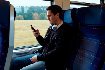 Caucasian young adult man sitting on train seat using smartphone with headphones around neck looking at screen with focused expression countryside landscape visible through window