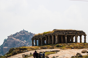 Visitors walk near ancient stone structure on hill with large rock formation in distance under clear sky