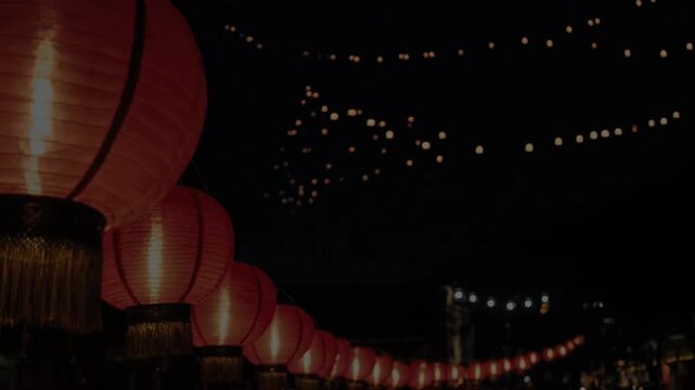 A row of glowing red lanterns on pillars at nighttime by the water with a bridge and string lights in the background for travel or festival promotions