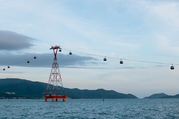 Obraz premium Cable car cabins suspended on steel cables above sea water with a red and white lattice support tower standing on yellow pillars against mountain view