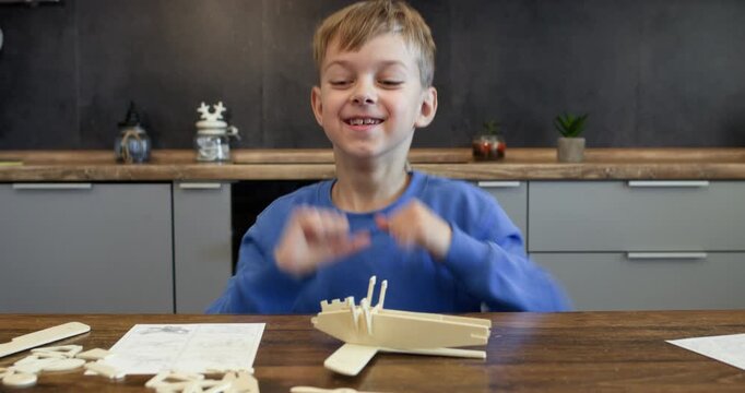 Joyful little boy spins incomplete wooden airplane model sitting at desk. Excited junior schoolboy plays with toy plane part made of plywood construction set in kitchen