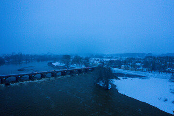 Aerial winter view of a historic arched bridge spanning a wide river, with snow-covered banks and rooftops under soft blue twilight, creating a tranquil and moody landscape.