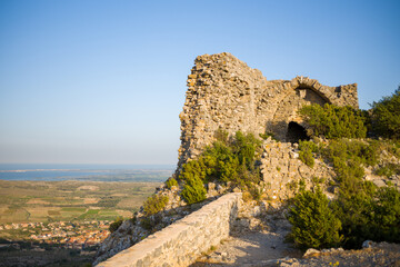 Ruined stone walls of Chateau de Opoul Perillos stand on a sunlit hillside, overlooking expansive green fields and a distant coastline under a clear blue sky.