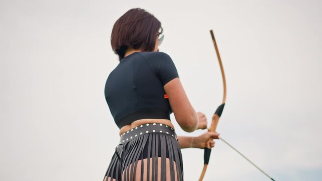 Full body archer raising bow arrow, fringe leather skirt and studded belt sway, wooden recurve bow pulled into aiming posture against cloudy sky, confident performance vibe with steady focus