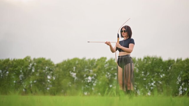 female archer standing in field, leather skirt and bow drawn under overcast sky tall grass and distant tree line frame solitary stance as she aims and releases arrow with focused, calm intensity,