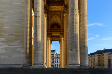 Massive neoclassical columns and ornate archway of the Pantheon in Paris, bathed in warm evening sunlight with city buildings framed in the distance.