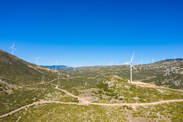 Multiple wind turbines line a rugged, sunlit mountain landscape near Thisbe, Greece, beneath a vibrant blue sky. Sparse vegetation and winding dirt roads highlight the remote, sustainable energy