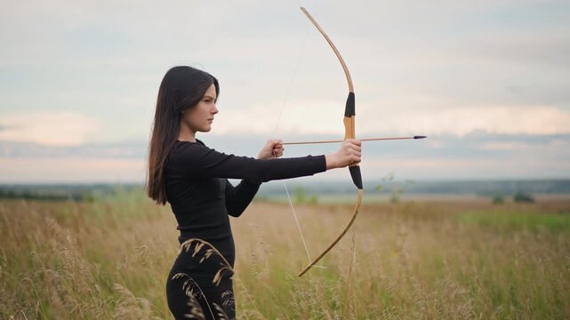 young woman archer in meadow drawing bow, aiming across grassy field at dusk. solitary warrior stance with leather bracer, arrow nocked and released, focused profile and steady gaze toward distant