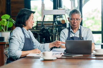 Senior asian couple cafe owners analyzing data and making decision using laptop in restaurant, showing cooperation and partnership in managing small family business successfully