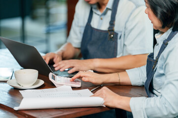 Asian elderly couple managing cafe finances calculating expenses with calculator laptop and notes on table, showing teamwork and collaboration in small family business management planning concept