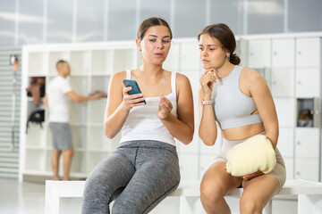 Positive young girl in casual sportswear holding smartphone and showing something to interested female friend while sitting together on bench in climbing gym locker area..