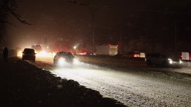 A wide shot captures a long line of cars with bright headlights stuck in a traffic jam on a snow-covered city road during a heavy night snowfall. Snowbanks line the roadside.