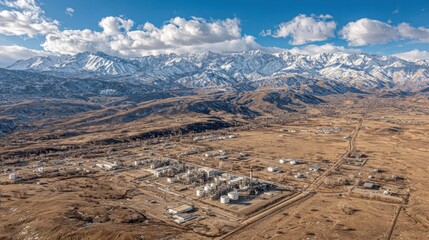 Aerial view of an industrial plant in dry terrain against snow-capped mountains. Suitable for themes on energy, infrastructure, industry, and environmental impact.