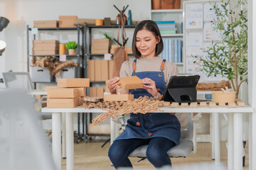 Professional Asian female entrepreneur in beige apron focused on sustainable packaging, checking eco-friendly products on tablet in creative workshop with recycled boxes and olive branches, green 