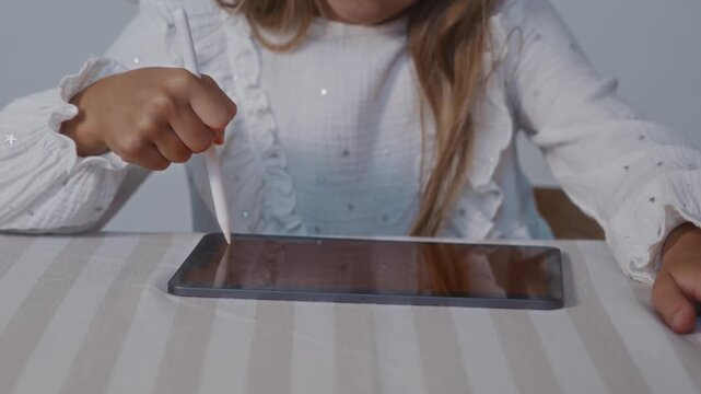 Medium close up shot from a slight top front angle shows a child using a stylus on a digital tablet placed on a table. The camera emphasizes hand coordination and learning through drawing. 4k footage