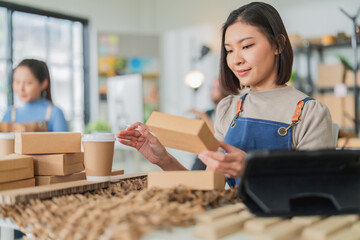 Professional Asian female entrepreneur in beige apron focused on sustainable packaging, checking eco-friendly products on tablet in creative workshop with recycled boxes and olive branches, green 