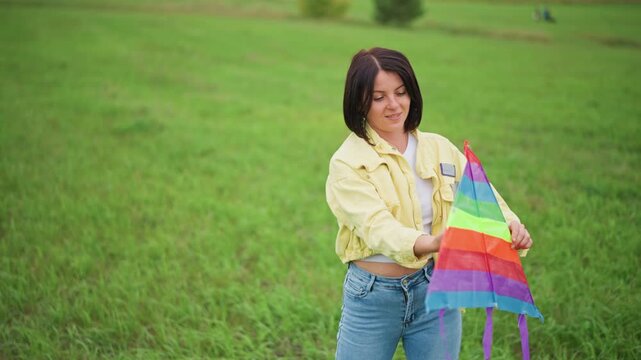 woman holding rainbow kite in field, calm overcast sky, yellow jacket and jeans, curly hair, gently testing kite before launch, slow deliberate movements, soft green meadow, peaceful solitary mood,