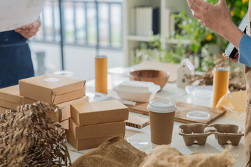Asian entrepreneurs team in sustainable workshop discussing eco-friendly biodegradable packaging, holding molded pulp trays and kraft boxes,creative green business startup collaboration bright room
